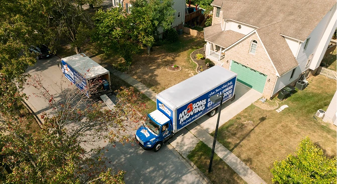 Aerial view of a professional moving truck and trailer parked outside a suburban home during a residential move. The image shows the moving vehicles positioned in the driveway and street, highlighting long-distance or out-of-state moving services in a neighborhood setting.