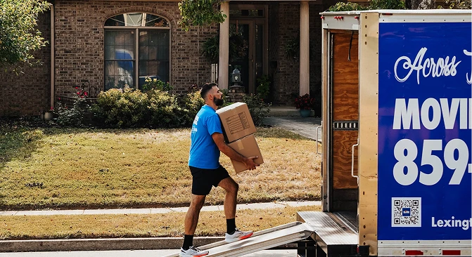 Professional mover carrying two cardboard boxes up a loading ramp into a moving truck parked in front of a suburban home. The image highlights residential moving services with a moving trailer, driveway, and front yard in the background.