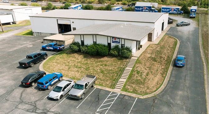 Aerial view of a professional moving and storage facility with warehouse buildings, office entrance, parking lot, company vehicles, and moving trucks. The image highlights a secure commercial storage location designed to support residential and business moving services.