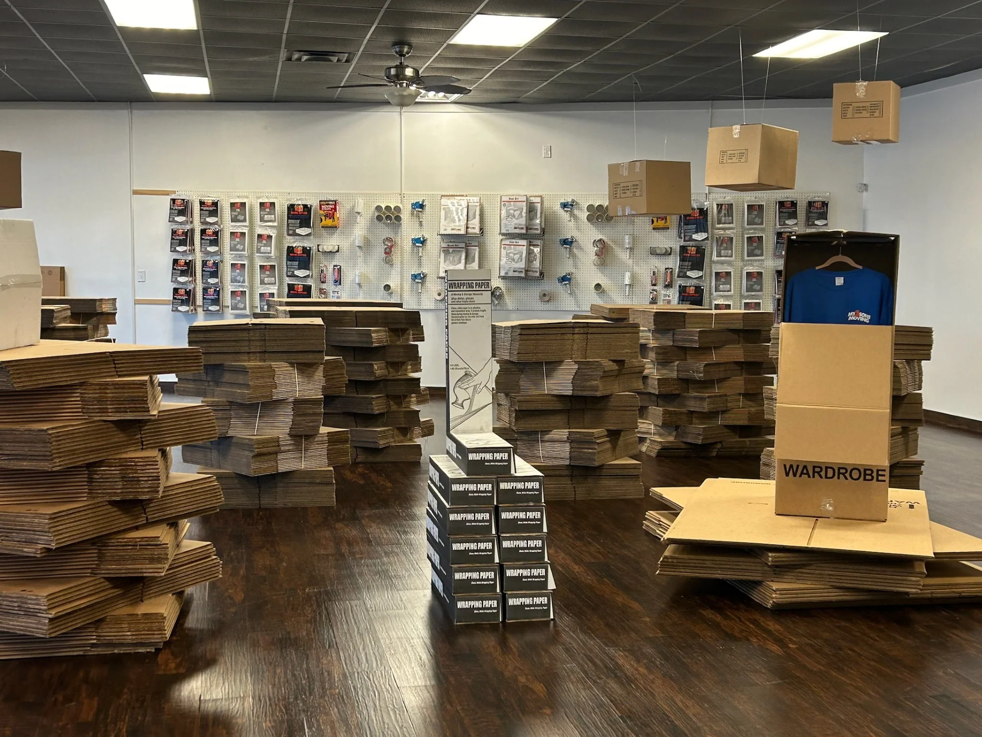 Moving supply store interior with stacks of cardboard boxes, wrapping paper, packing supplies, and a wardrobe box display.